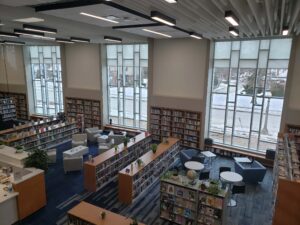 Interior of a spacious library with large windows, bookshelves, seating areas, and blue carpet.